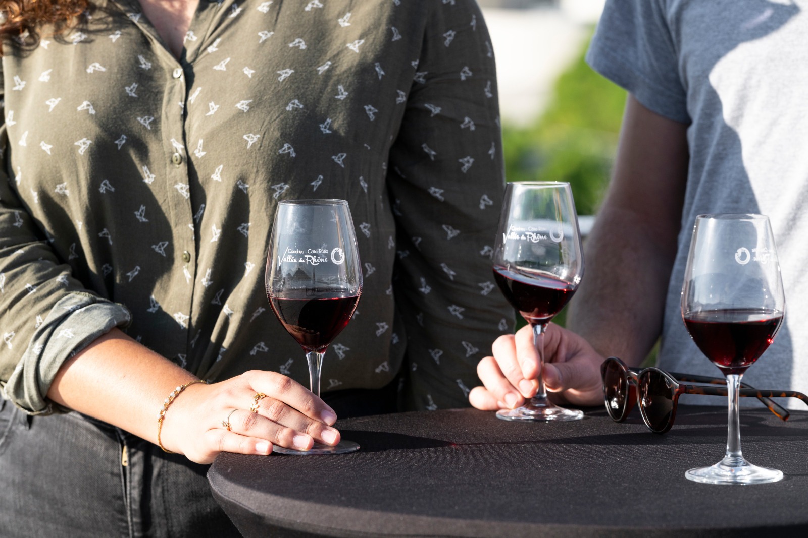 3 verres de vin posés sur un table. Deux personnes se tiennent sur la photo et tiennent chacun un verre dans la main.