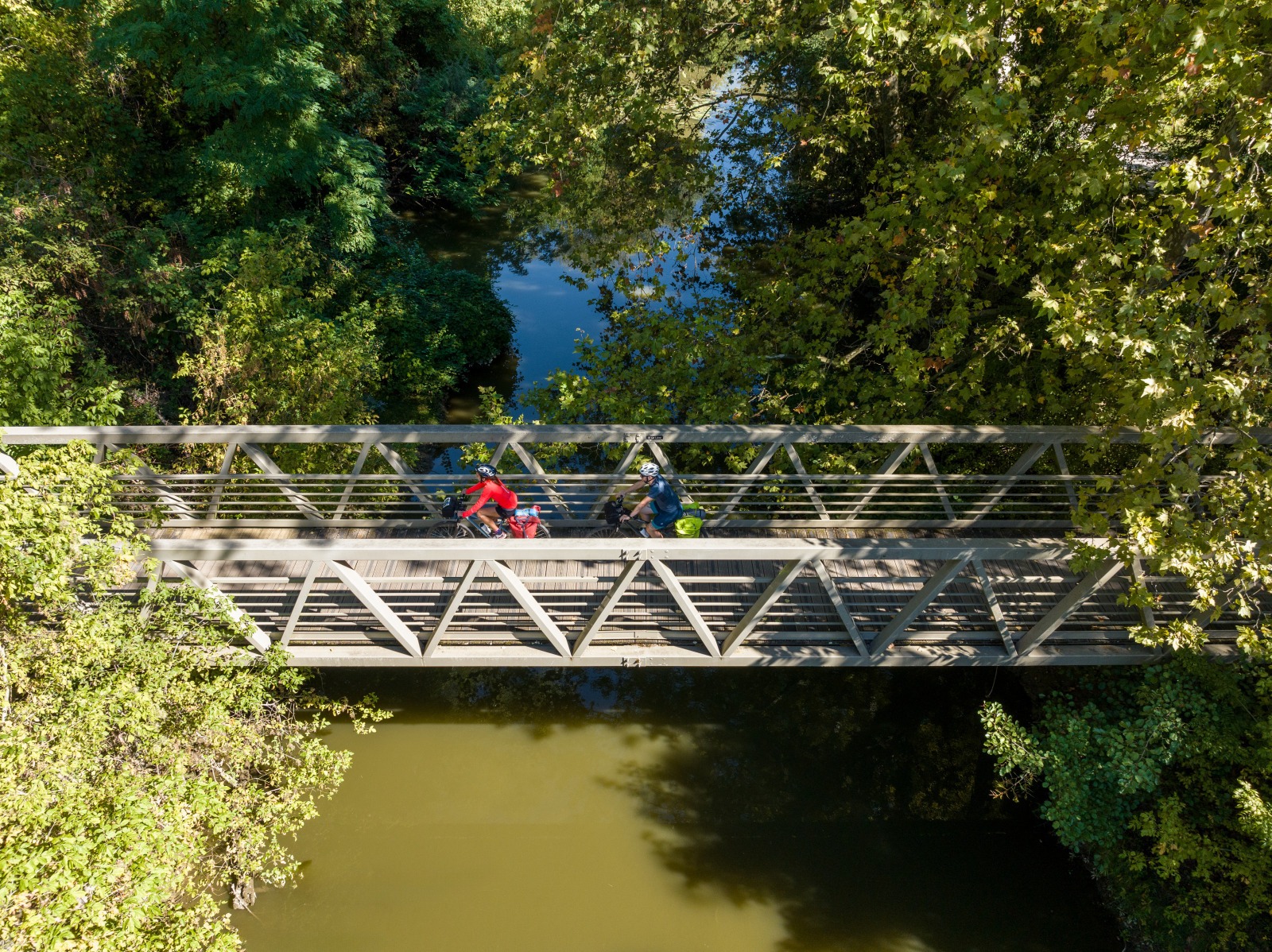 Dos personas en bicicleta cruzan un pequeño puente peatonal rodeado de vegetación y agua.