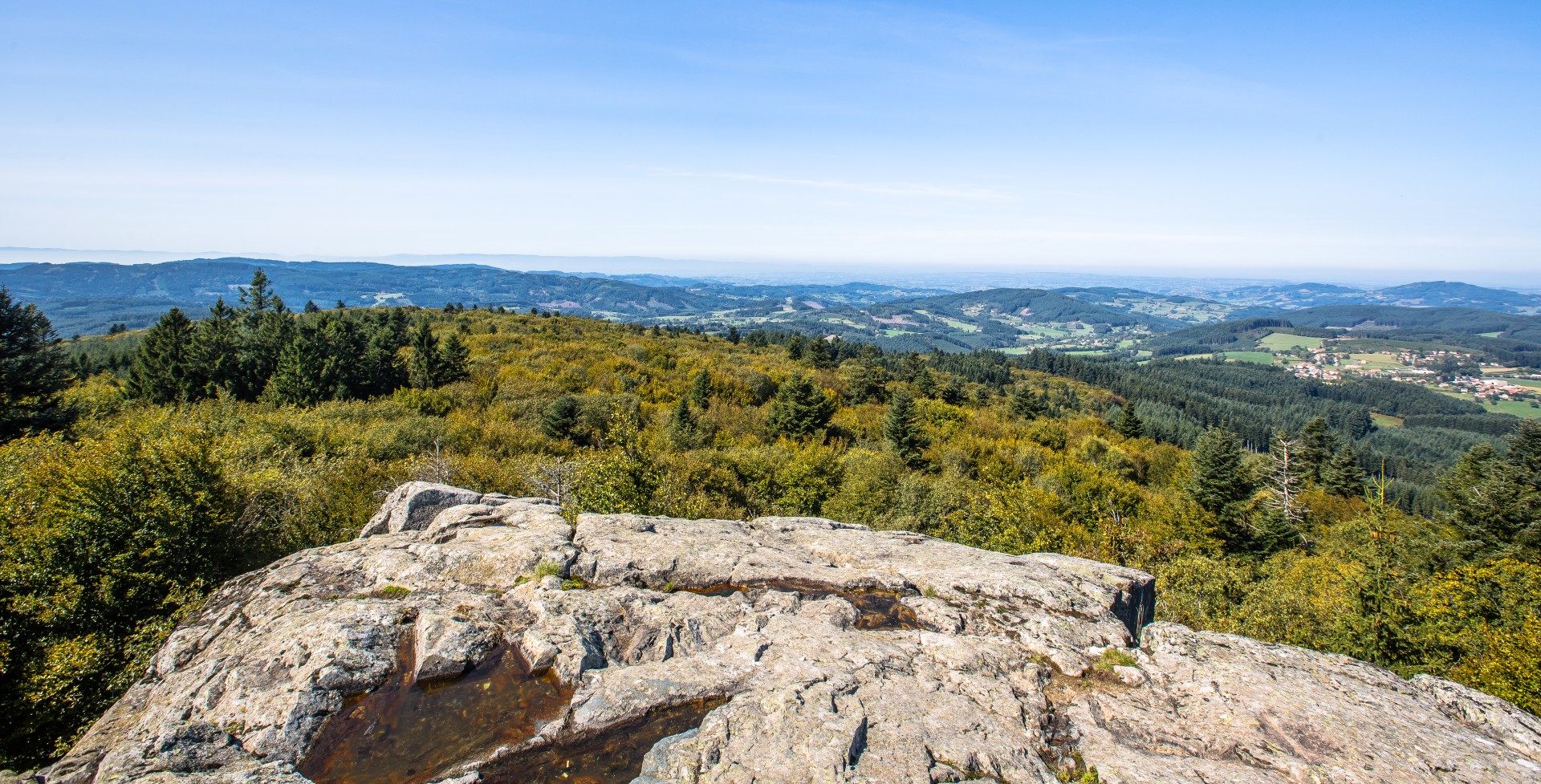 Promontoire rocheux avec vue panoramique sur les collines forestières du Beaujolais Vert et sous un ciel bleu dégagé. Site de la roche d'Ajoux