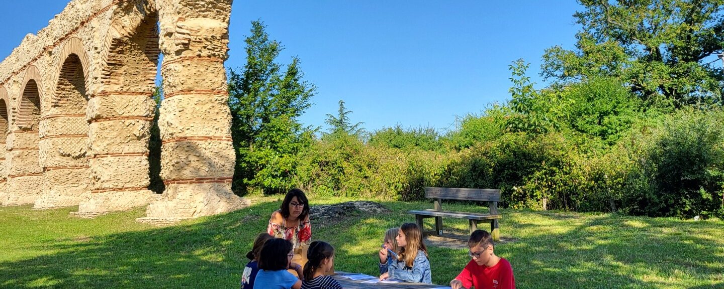 Une table de pique-nique en premier plan avec une guide qui explique aux 6 enfants assis sur la table de pique-nique l'histoire de l'aqueduc du Giers à Chaponost dans les monts du lyonnais, il situé en arrière-plan de la photo. La nature est verdoyante, il fait soleil.