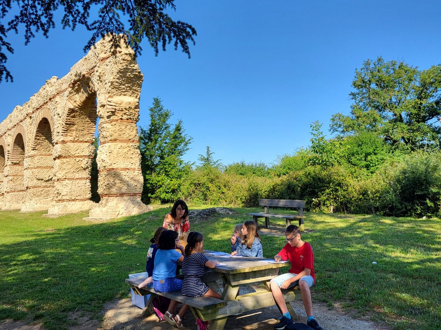 Une table de pique-nique en premier plan avec une guide qui explique aux 6 enfants assis sur la table de pique-nique l'histoire de l'aqueduc du Giers à Chaponost dans les monts du lyonnais, il situé en arrière-plan de la photo. La nature est verdoyante, il fait soleil.