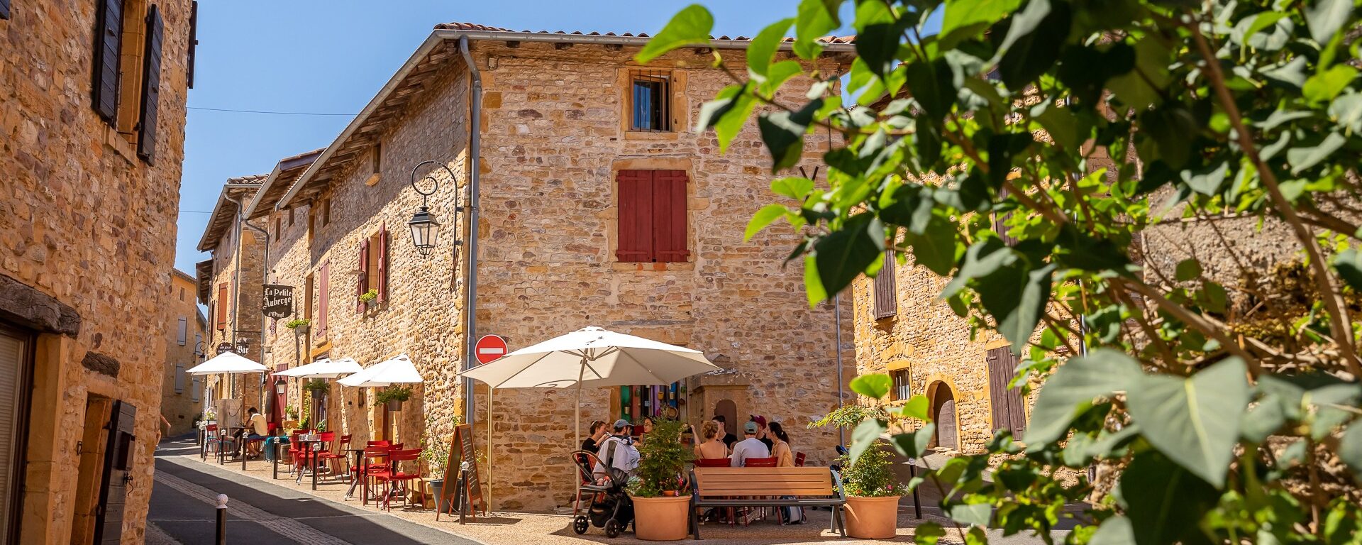 Village d'Oingt sous le soleil estival. Rue principale bordée par une terrasse de café. Des touristes sont installés autour de tables rouges sous des parasols blanc. Beaujolais des Pierres Dorées